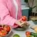 Person in pink overshirt holding a fresh tomato with various organic vegetables on a countertop.